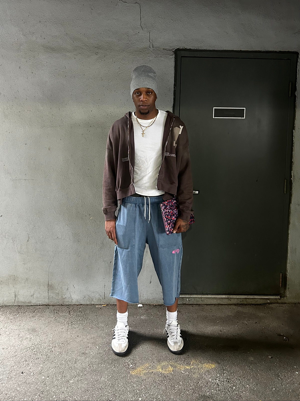 A person stands in a concrete room wearing a gray beanie, brown zip-up hoodie, white t-shirt, light blue cropped sweatpants, white sneakers, and holding a small patterned pouch.
