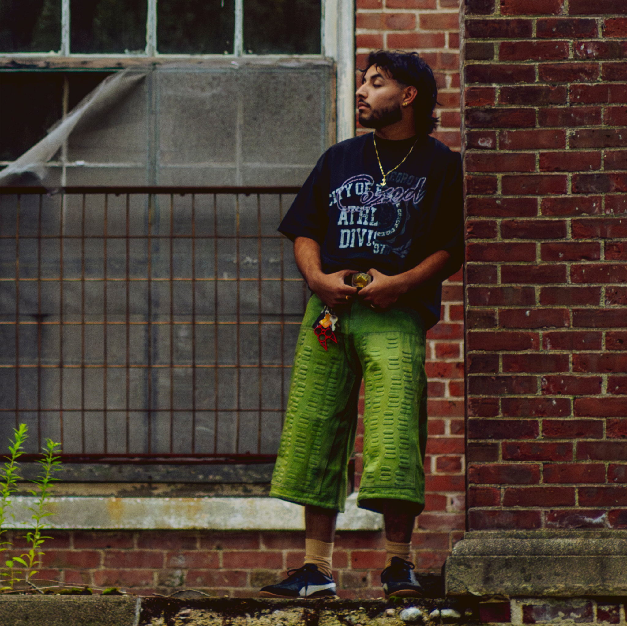 Person wearing a black t-shirt and green shorts standing against a brick wall.
